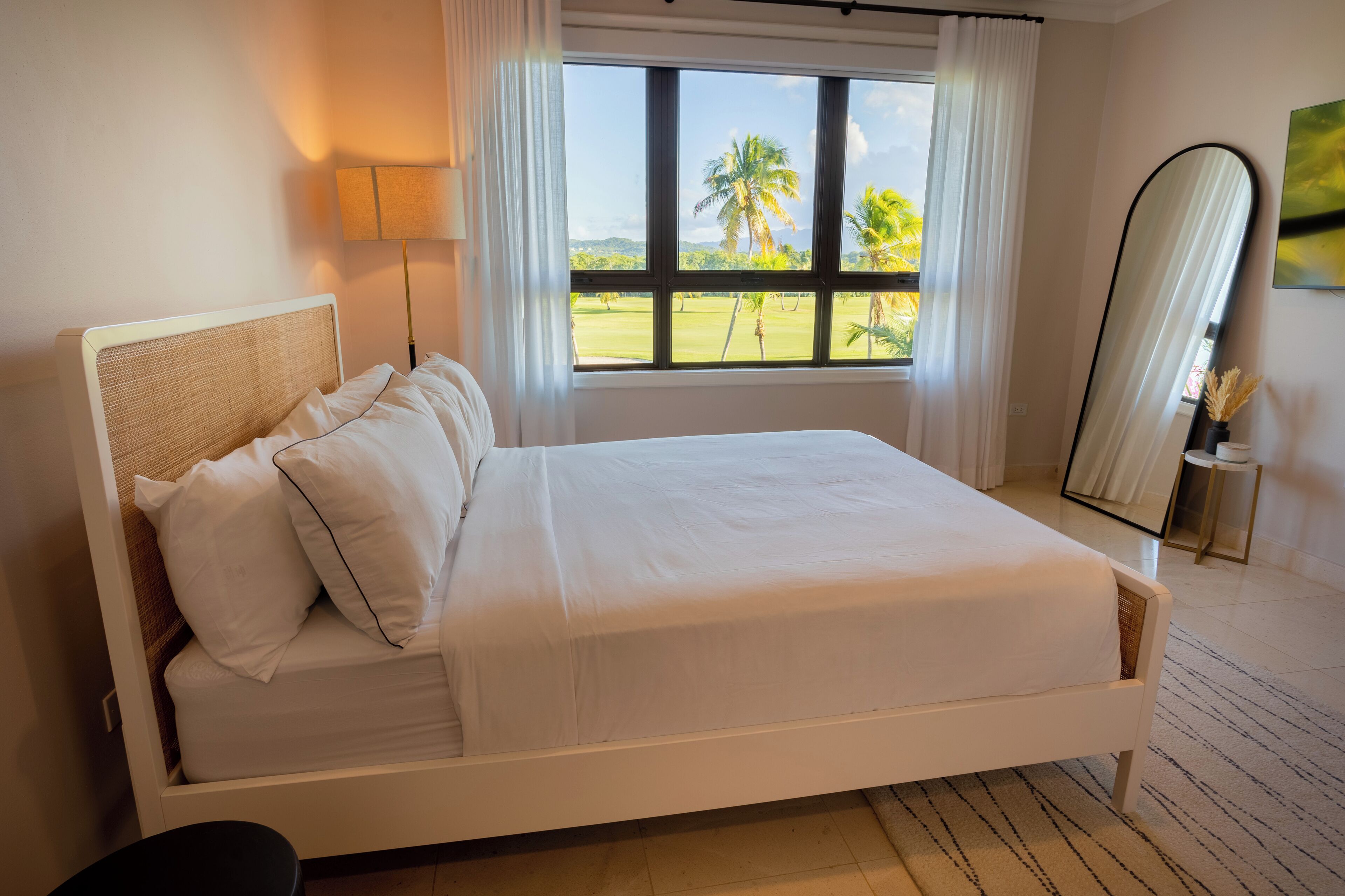 Een keurig opgemaakt bed met witte lakens, een groot raam met uitzicht op palmbomen en een staande spiegel aan de muur.