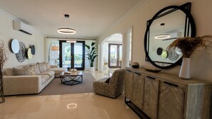 Ein geräumiges Wohnzimmer mit Sofa, Ohrensessel und Couchtisch, einem großen Spiegel und Blick ins Freie.