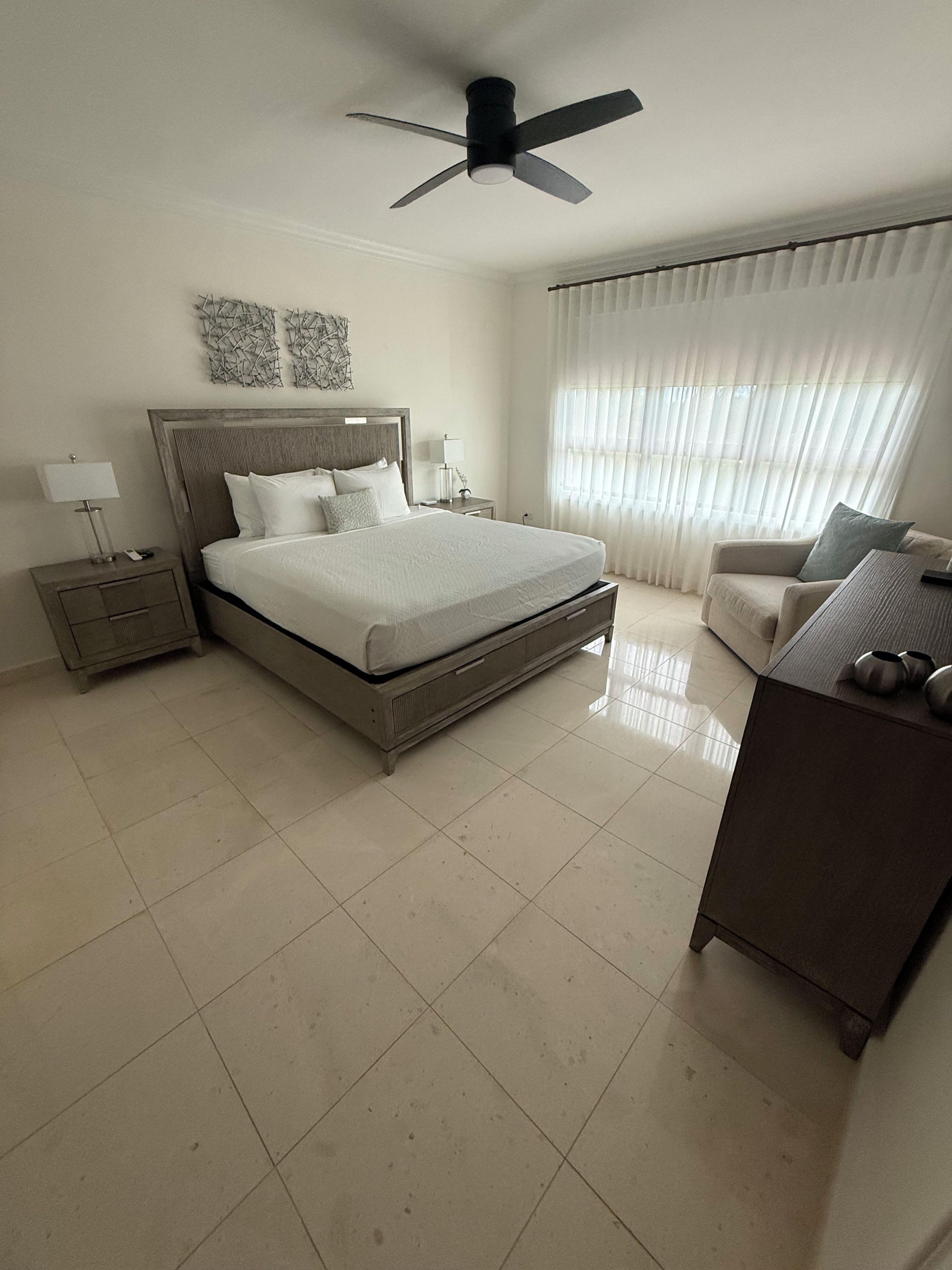 A hotel room with a large bed, a desk, a chair, a ceiling fan, and a window with curtains.