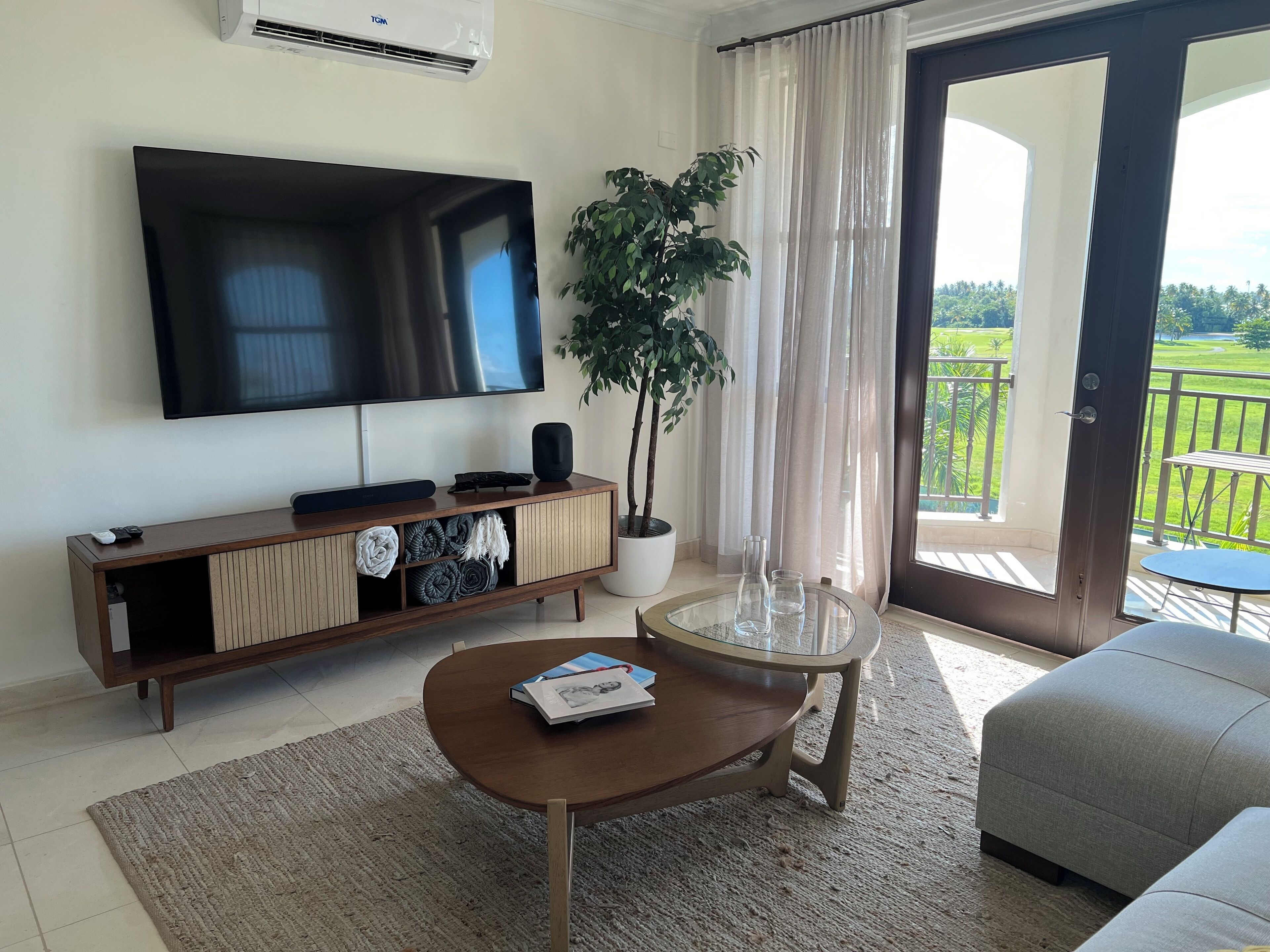 Uma sala de estar com televisão de tela plana, um vaso de planta, uma mesa de vidro com um livro e uma varanda com vista.