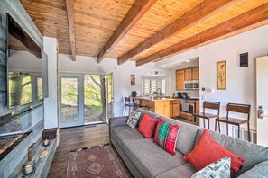 Un salón acogedor con un sofá gris, vigas de madera en el techo y un amplio ventanal con vistas a un jardín.