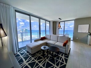 A modern living room with a large sectional sofa, a coffee table, and a view of the city and ocean through floor-to-ceiling windows.