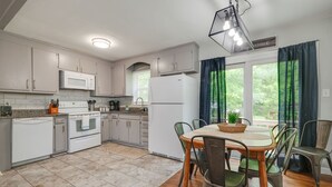 Una cucina moderna con elettrodomestici bianchi, un'area pranzo con tavolo in legno e sedie in metallo, e vista sul verde dalla finestra.