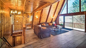 A cozy cabin interior with wooden walls, a loft-style ceiling, and a large window offering a view of trees. - Running Springs Cabin w/ Large Deck + View! (Running Springs)