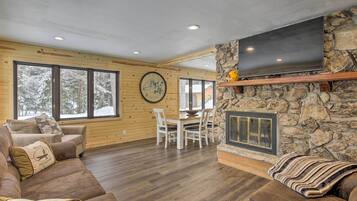 A cozy living room with a stone fireplace, wooden walls, and a dining area with a large window overlooking a snowy landscape.