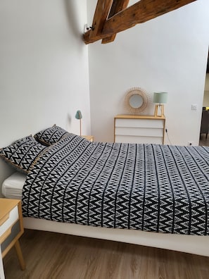 A bed with a patterned bedspread, a bedside table with a lamp, a small round mirror, and a wooden headboard.