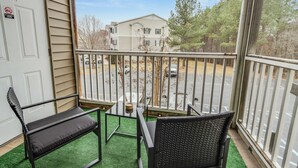 Un balcon avec deux chaises noires, une petite table et une vue sur un parking et des immeubles.