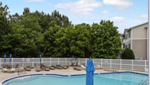 Une piscine entourée d’une clôture blanche et d’arbres, avec des chaises longues et des parasols à proximité.
