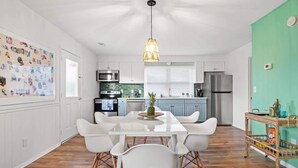 A modern kitchen with white cabinets, a stainless steel refrigerator, and a dining area with a white table and chairs. - Beach Nest 4 Stylish 2Min Walk to Beach (North Myrtle Beach)