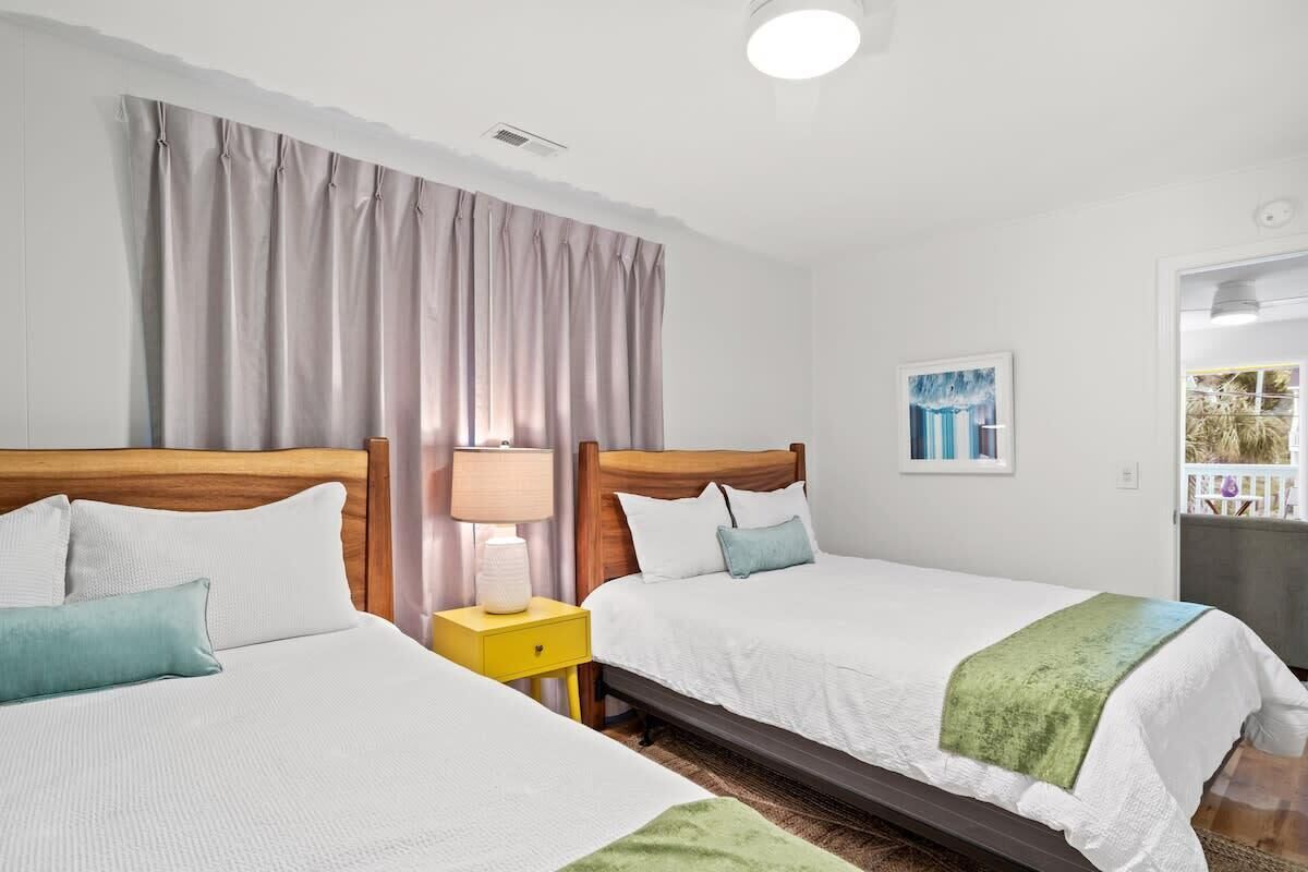 A hotel room with two beds, a wooden headboard, a nightstand with a lamp, a painting on the wall, and a view of the outside balcony.