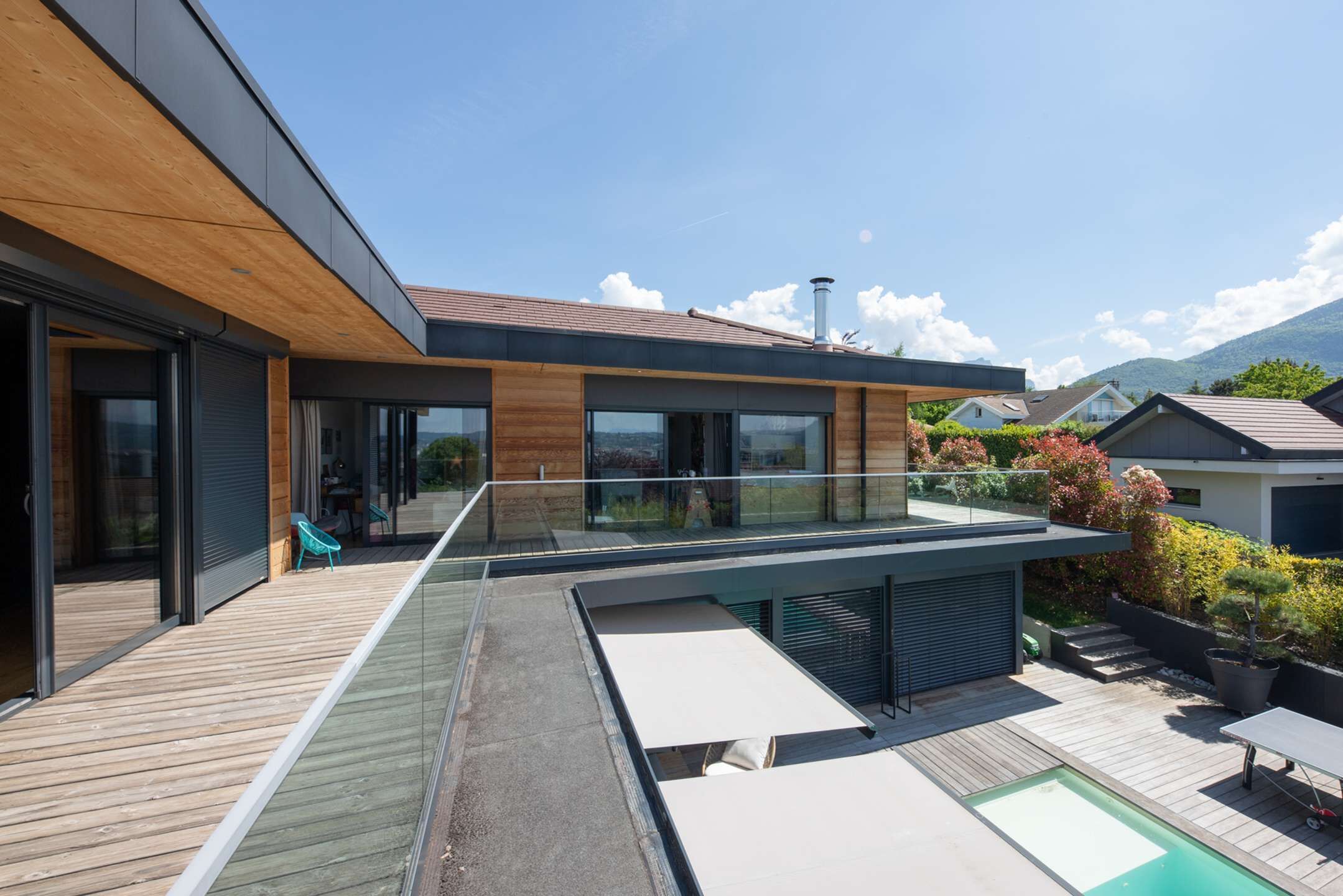 Una casa moderna con terraza de madera, piscina y balcón con vistas a un jardín.