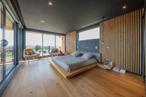 Ein modernes Schlafzimmer mit einem großen Bett, einem Sessel und Blick durch große Fenster.