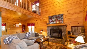 A cozy cabin interior with a fireplace, wooden walls, a sofa, and a television mounted above the fireplace. - Rustic Cabin With Mountain Views (Todd)