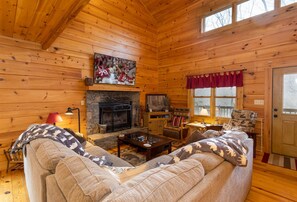 A cozy log cabin interior with a fireplace, a television mounted above it, and a comfortable seating area with a sofa and armchairs.
