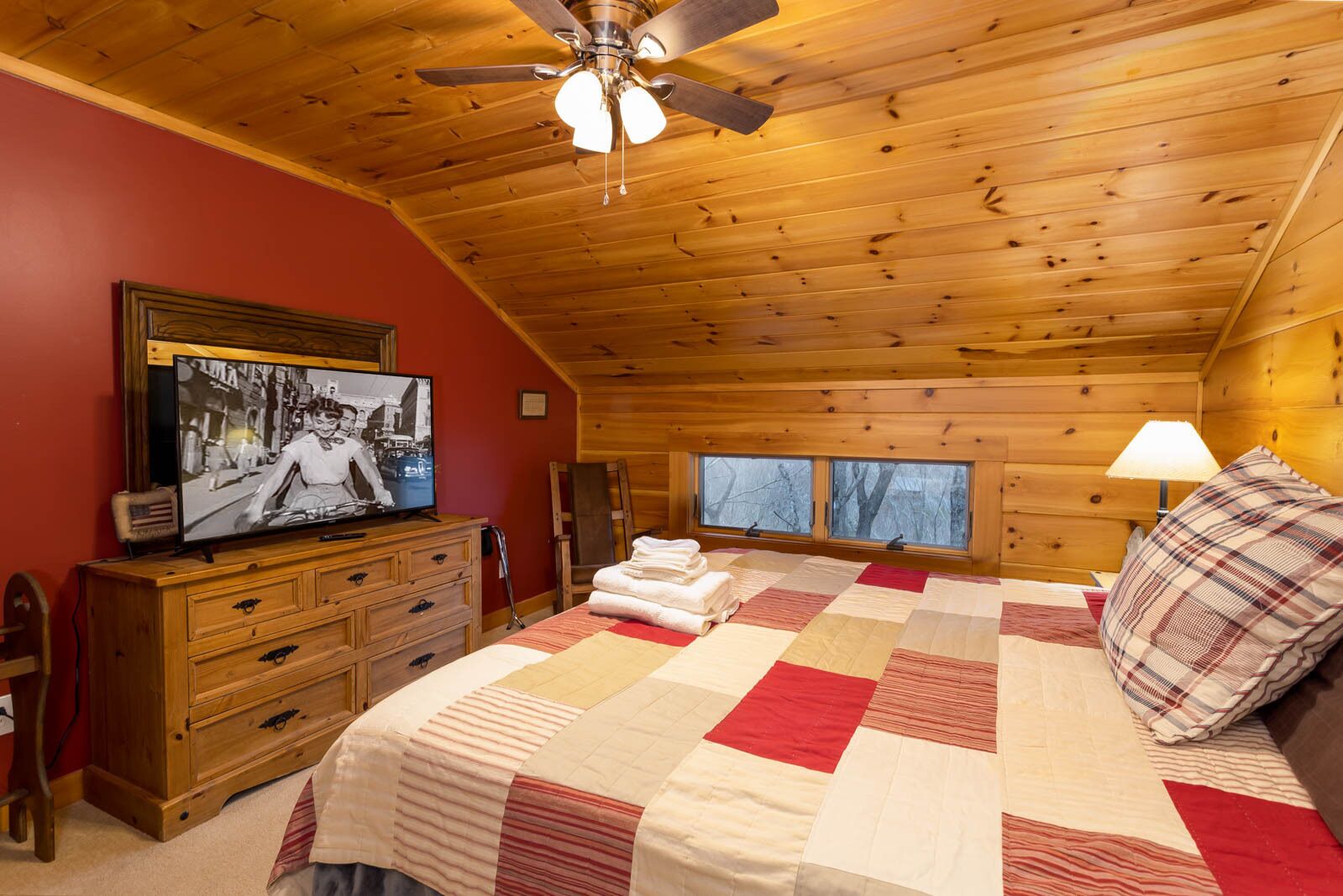 Une chambre confortable dans un chalet en rondins, avec un plafond en bois, un lit recouvert d’une courtepointe, un téléviseur posé sur une commode en bois et une fenêtre donnant sur la forêt.