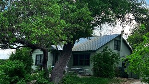 A rustic house with a metal roof, surrounded by lush greenery and a gravel path.