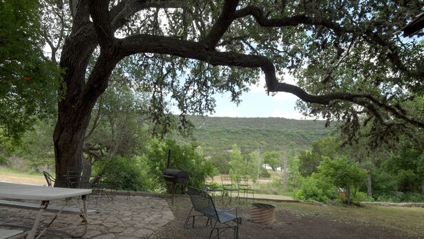 A picnic area with stone tables and metal chairs, surrounded by trees and a view of a hilly landscape.