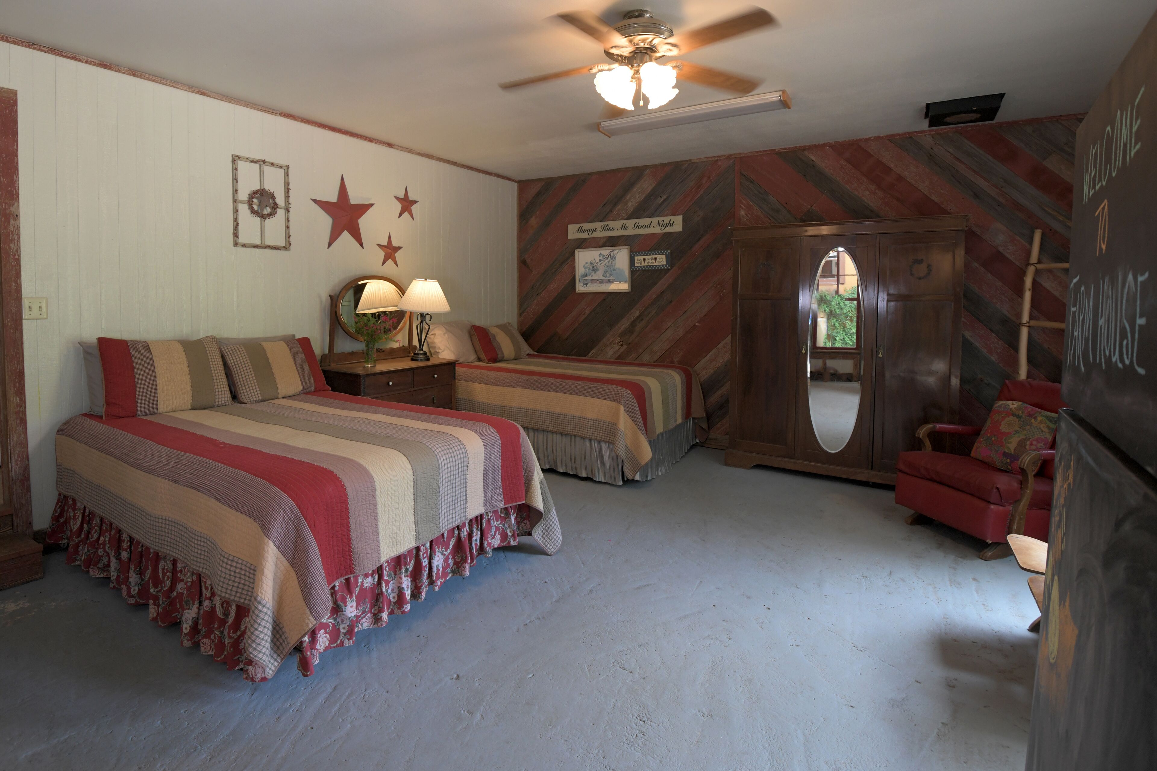 A bedroom with two beds, a red armchair, a wooden wardrobe, and a ceiling fan.