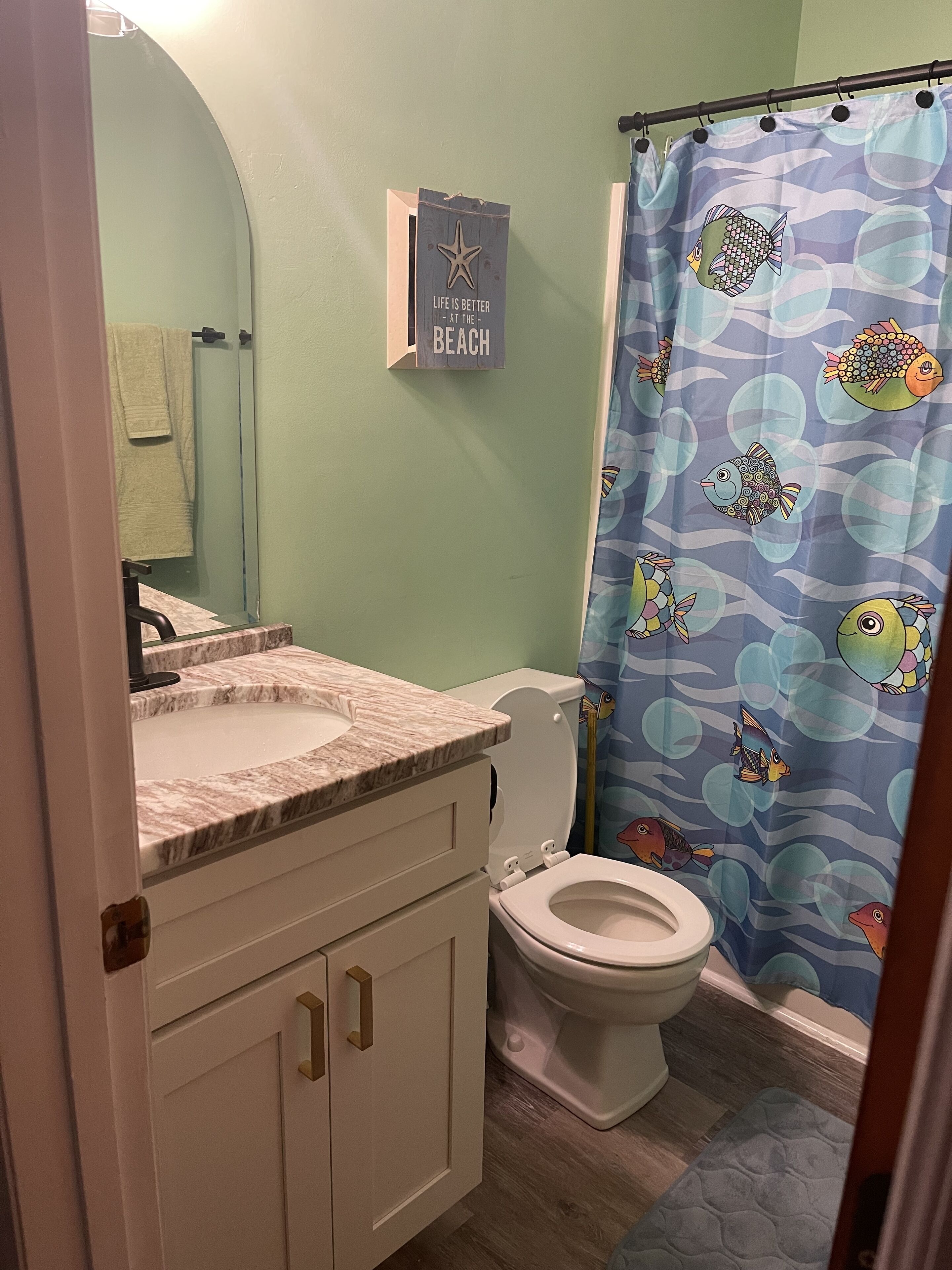 A bathroom with a toilet, sink, and vanity. There is a shower curtain with a fish pattern. A sign on the wall reads "Life is better in the beach."