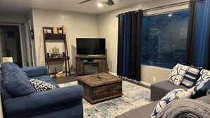 Una sala de estar moderna con un sofá, un reposapiés, una mesa de centro, televisor, estantería y un gran ventanal con cortinas.