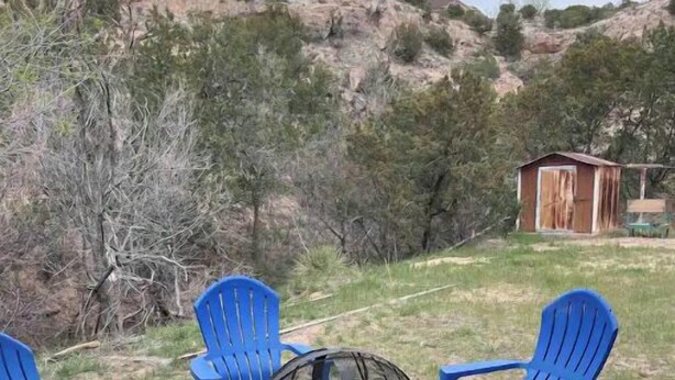 Un área de estar con sillas de plástico azul y un brasero central, rodeada de un paisaje herboso con una pequeña estructura de madera al fondo.