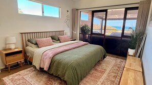 Quarto com cama, mesa de cabeceira, abajur e vista para o exterior por uma porta de correr.