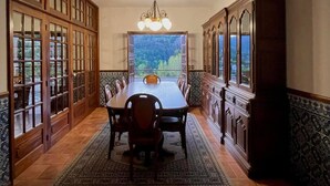 Une salle à manger avec une longue table, des chaises, un lustre et de grandes fenêtres donnant sur un paysage verdoyant.