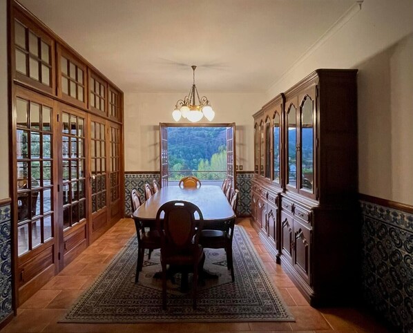 A dining room with a long table, chairs, a chandelier, and large windows with a view of greenery.