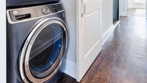 A laundry area with two front-loading washing machines, a white door, and a hallway leading to a bright room.