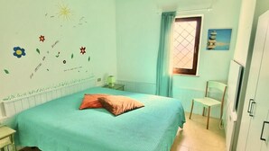 A bedroom with a teal bedspread, a single orange pillow, a nightstand, a chair, a window with curtains, and a wall with decorative elements. - Sa Chessa House Two-room apartment in San Teodoro (San Teodoro)