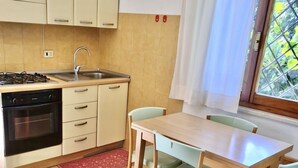 A compact kitchen with a gas stove, oven, sink, and light-colored cabinets. There is a dining table with chairs and a window with a curtain. - Sa Chessa House Two-room apartment in San Teodoro (San Teodoro)