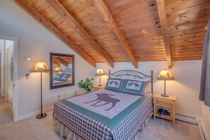 Une chambre avec un plafond en bois, un lit recouvert d’une courtepointe à motifs d’élans, deux lampes de chevet, une table de chevet avec une lampe et un miroir sur le mur.