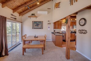 Un grand espace à manger avec une table et des chaises en bois, une grande fenêtre offrant une belle vue et un coin cuisine visible à travers une porte ouverte.