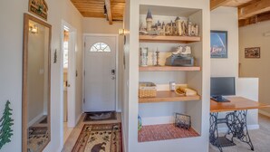 Un pasillo con techo de madera, un cartel de bienvenida, una puerta con ventanal, estantes con diversos objetos, una mesa con computadora y una máquina de coser.