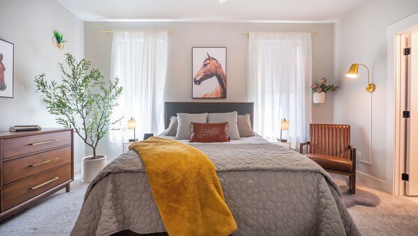 Un dormitorio con cama, dos lámparas de noche, una silla de madera, un aparador, una planta en maceta y un cuadro en la pared.
