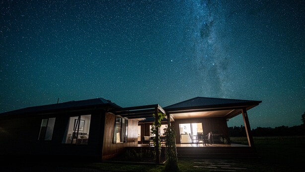 A modern house with large windows and a covered patio, under a starry night sky.