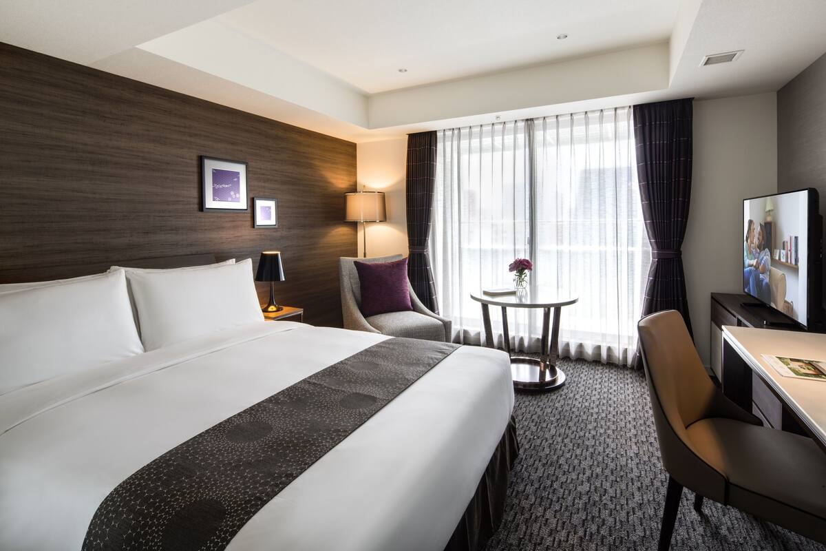 A hotel room with a large bed, a desk, a chair, a TV, and a window with curtains.