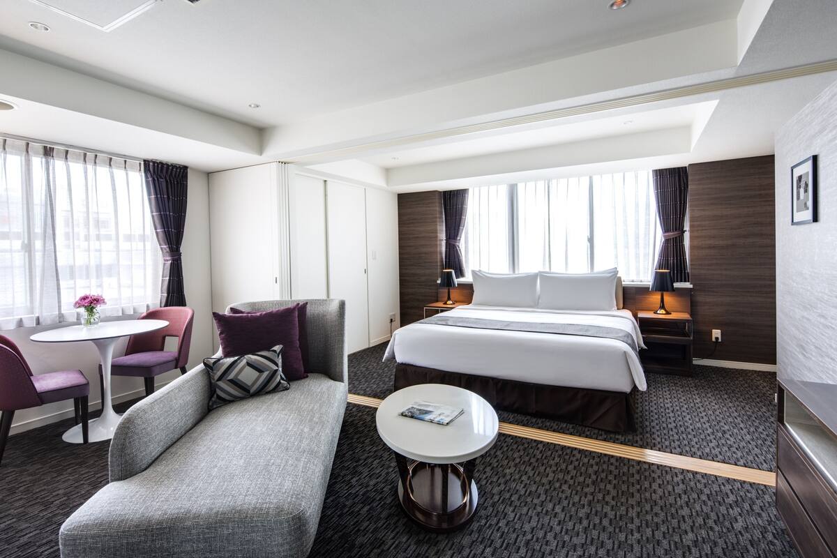 A modern hotel room with a large bed, a sofa, a small table, and a window with curtains.