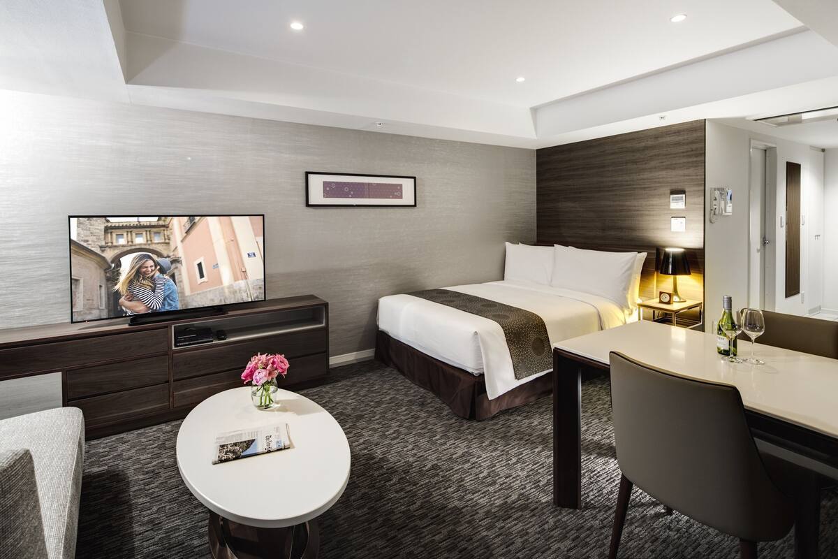 A modern hotel room with a bed, a desk, a TV, and a small table with a vase of flowers.