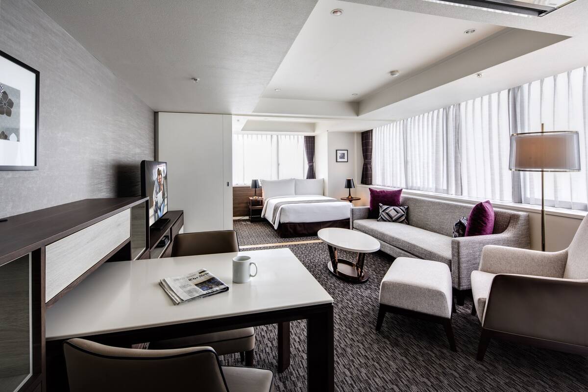 A modern hotel room with a bed, sofa, armchair, coffee table, and a desk with a chair.