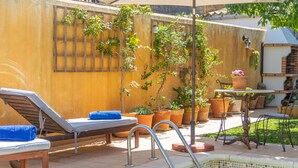 Área à beira da piscina com espreguiçadeiras, uma mesa pequena e plantas em vasos.