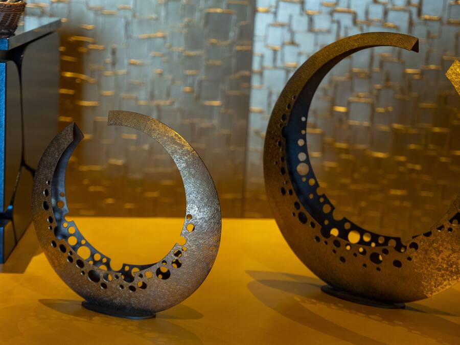 Two metallic crescent sculptures with holes, placed on a table against a patterned wall.