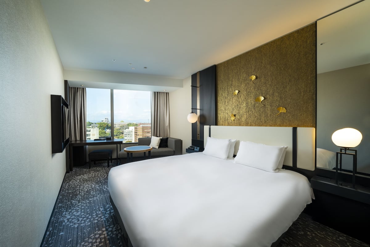 Hotelkamer met een groot bed, een bureau, een stoel, een kleine tafel en een groot raam met uitzicht over de stad.