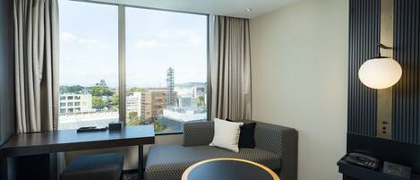 Ein Hotelzimmer mit großem Fenster, einer Couch, einem kleinen Tisch und einem Schreibtisch.