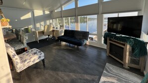 A spacious living room with a large window overlooking a body of water, a dark-colored sofa, a small table, and a television on a wooden stand.