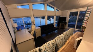 A cozy living room with a sofa, a small table, and a TV. Large windows offer a view of the outdoors.