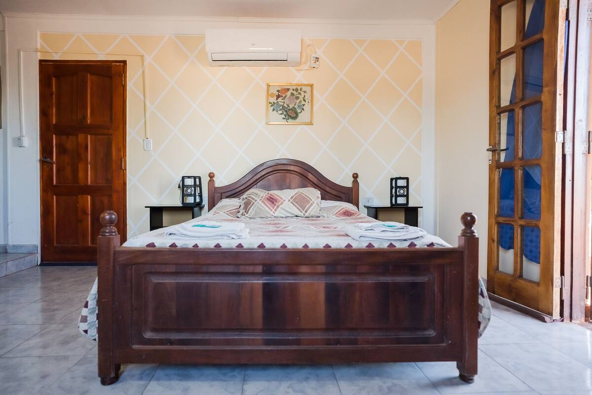 Une chambre à coucher avec un lit en bois, un climatiseur fixé au mur, une porte et une fenêtre avec un rideau bleu.