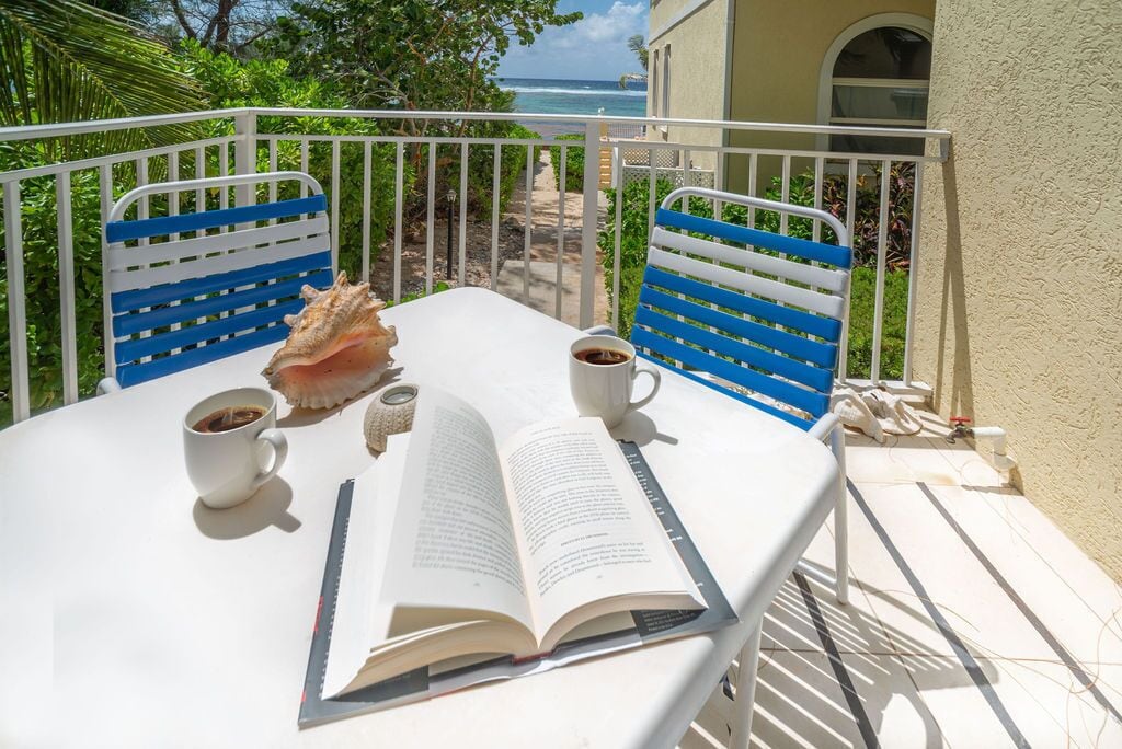 Una mesa con dos sillas, un libro y dos tazas de café en un balcón con vista al océano.