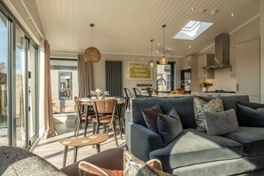 A modern kitchen with a skylight, dining area with a table and chairs, and a living room with a sofa and cushions.