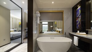 A modern hotel bathroom with a large freestanding bathtub, a mirror, and a view of a bedroom with a bed and a window.